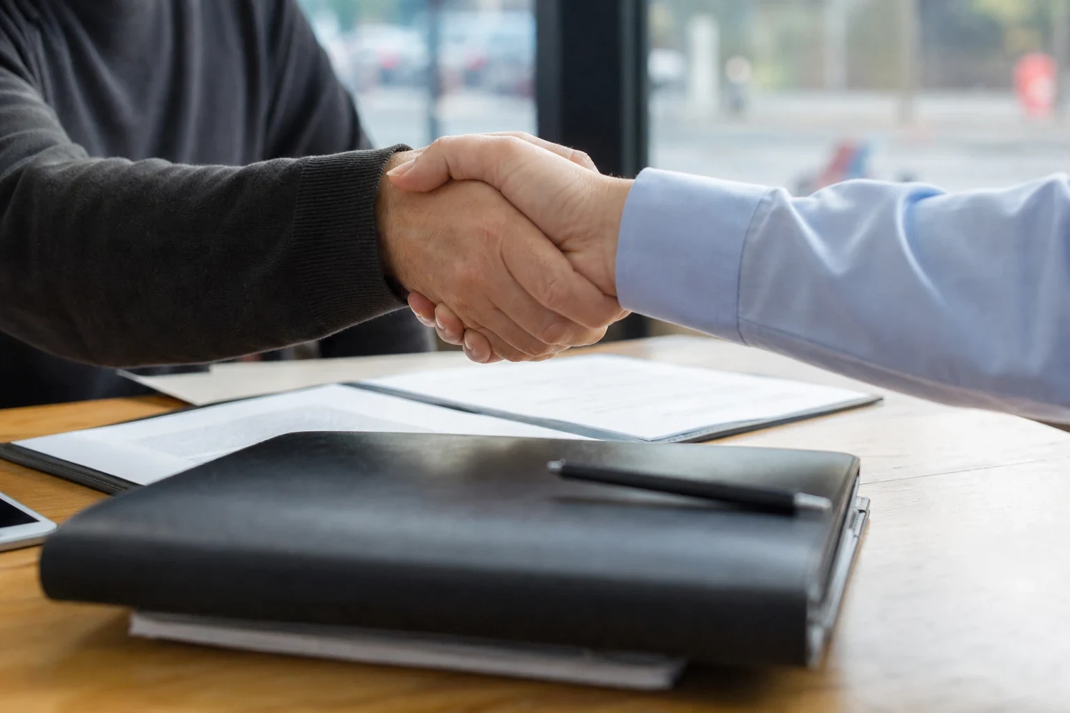Close-up of a handshake over a wooden desk between a person in a dark sweater and a person in a light blue shirt, with a leather folder, pen and papers