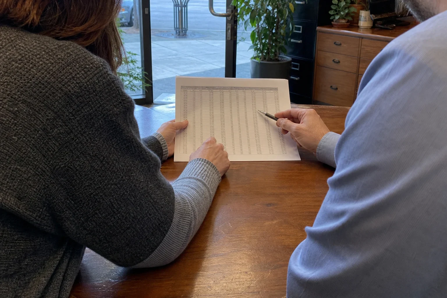 Two people at a wooden table in a storefront office review a printed page of figures, one pointing with a pen, near a glass door, filing cabinets and plants