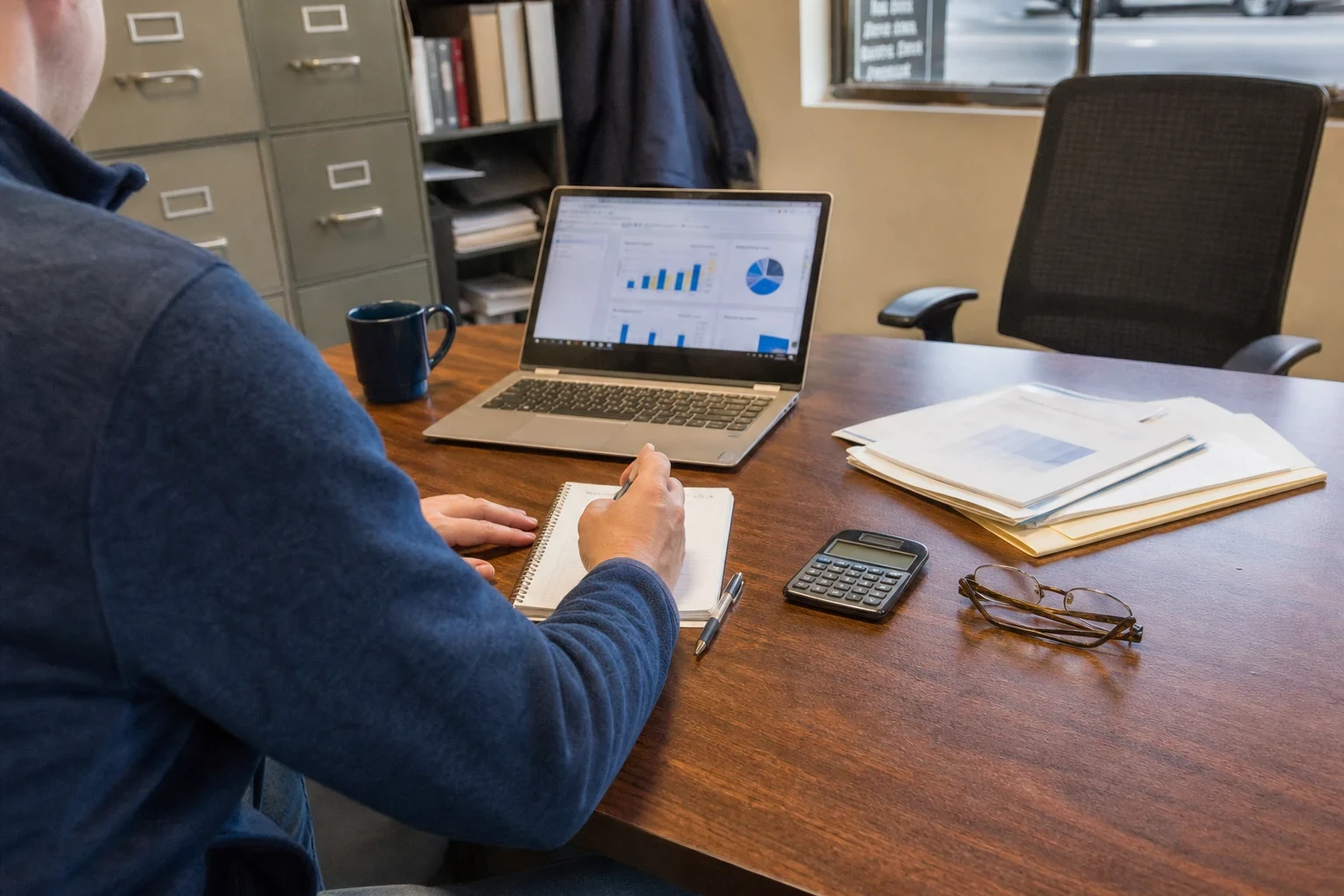 Man in a navy sweater writes notes at a wooden desk beside a laptop showing charts, a calculator, reading glasses and paper files in a small office with filing cabinets