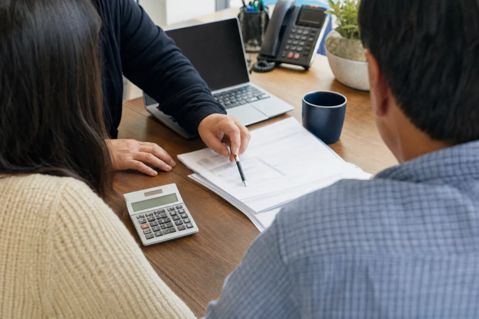 Over-the-shoulder view of two clients at a wooden desk as an agent points a pen at papers beside a laptop, calculator, desk phone, mug and potted plant